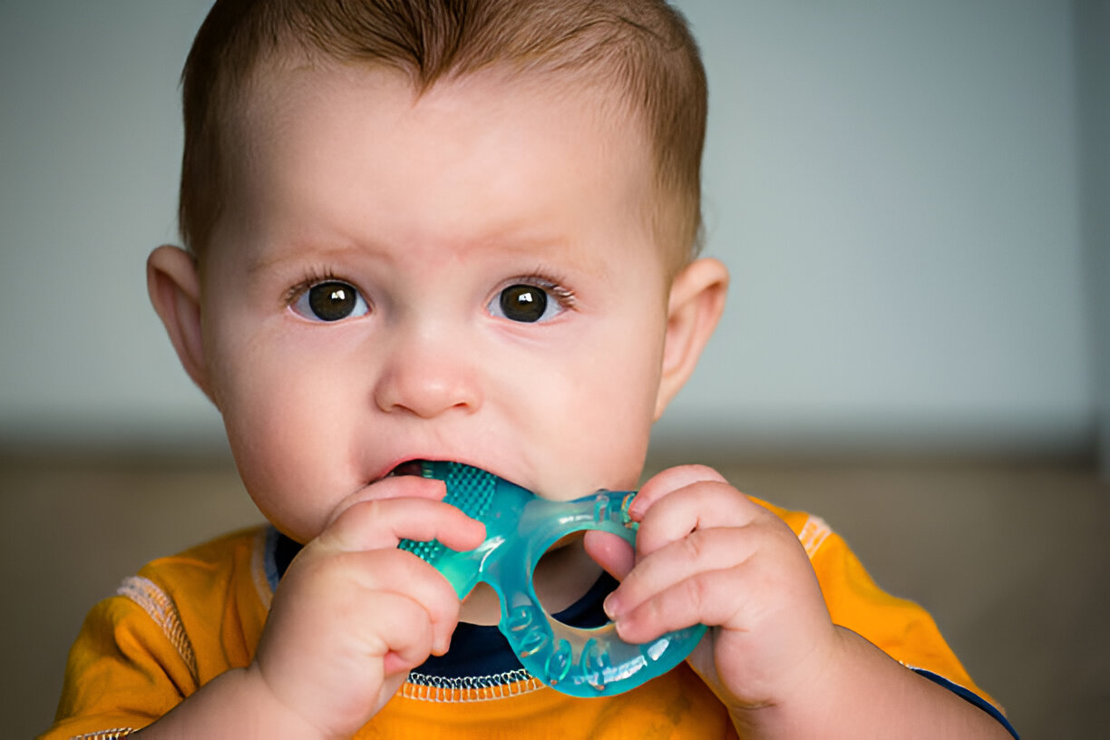 Teething baby chewing on a blue teething toy while wearing an orange shirt, providing relief from teething discomfort.