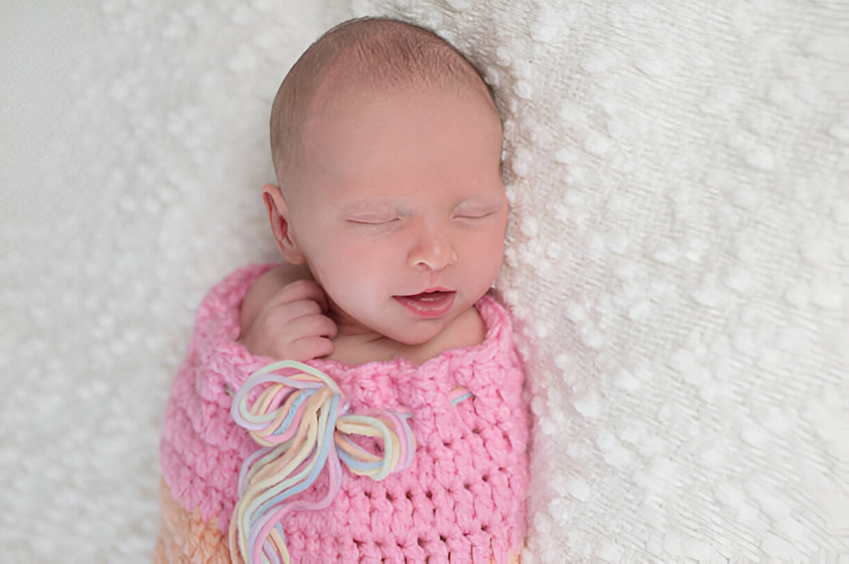 Newborn baby peacefully sleeping in a pink crochet sleep sack with a pastel rainbow bow, resting on a white textured blanket.