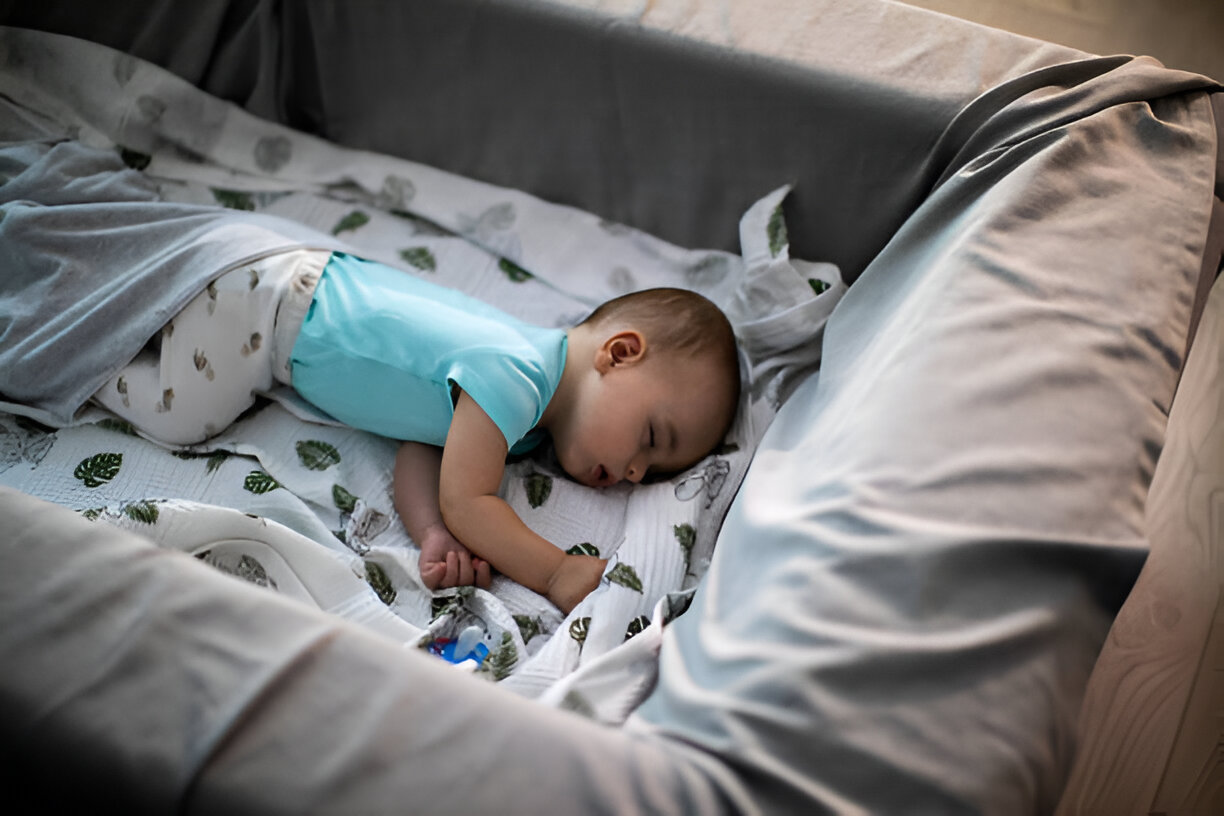 A baby peacefully sleeping in a crib, dressed in a blue onesie, lying on a white blanket with leaf patterns. Despite the serene moment, the image reflects the common theme of Babies Fight Sleep, showing a baby finally at rest after likely resisting sleep.