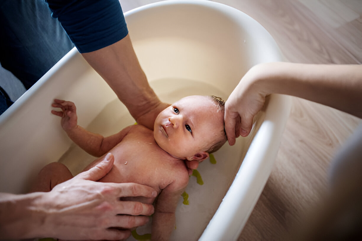 A newborns baby being gently bathed in a small tub by two adults, with one holding the baby’s head for support while the other washes the body, ensuring a safe and comfortable bath time for the infant.